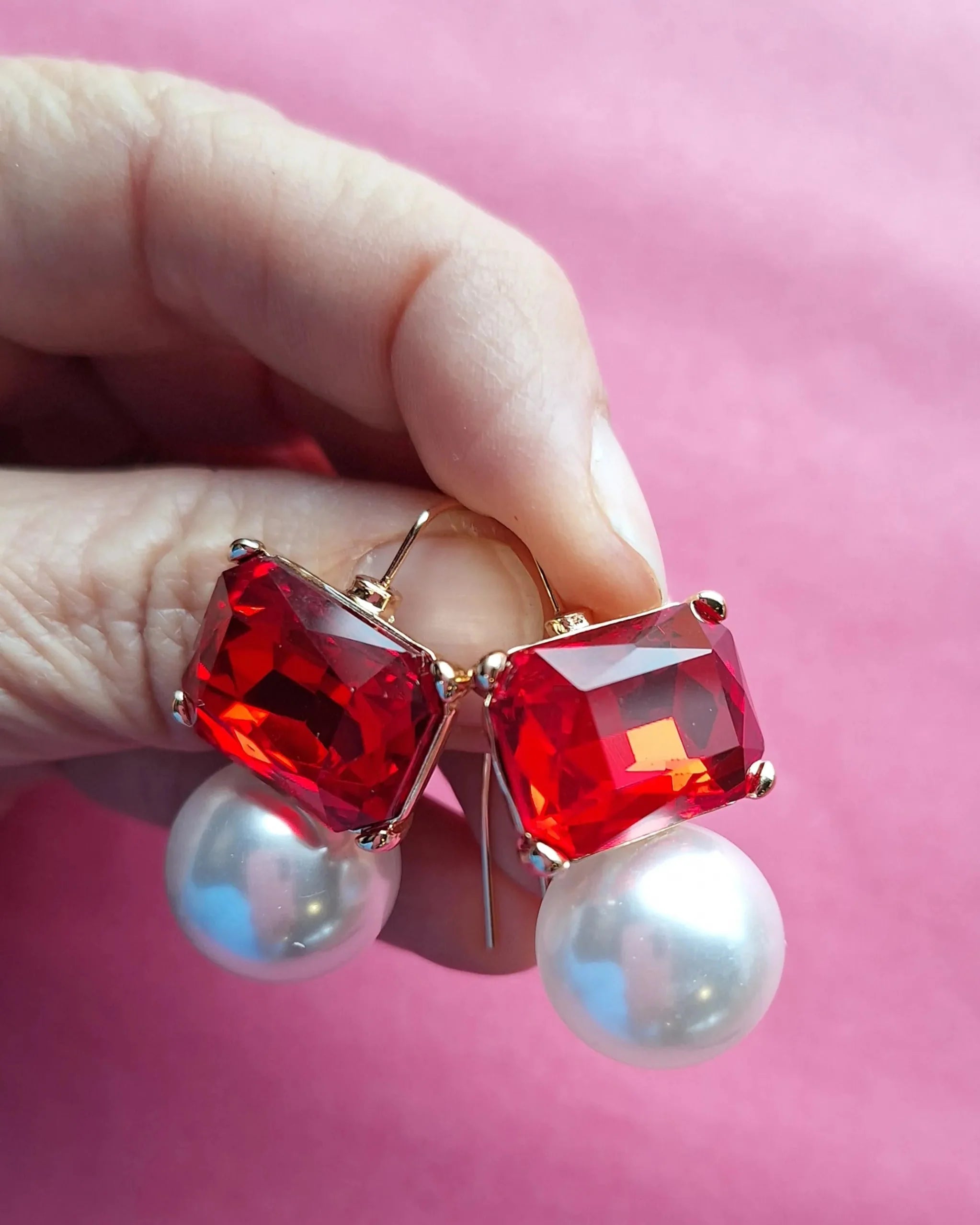 Earrings with red gemstones and pearls held by a hand against a pink background