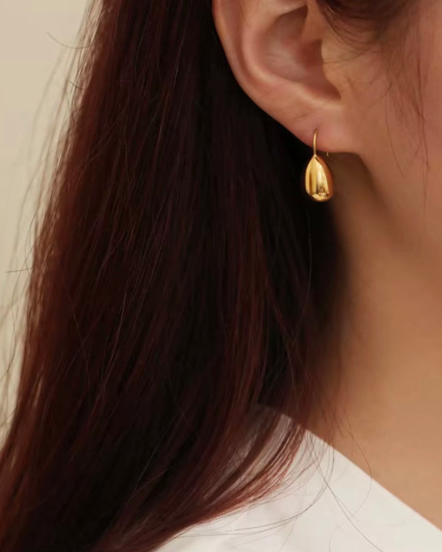 A close-up of a woman's ear wearing a small gold teardrop-shaped dangle earring, with dark brown hair and a white top visible against a soft beige background.