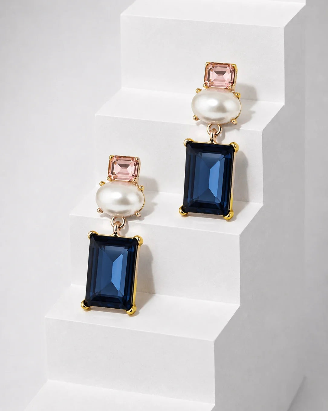 A pair of gold-tone drop earrings with rectangular navy blue gemstones, round white faux pearls, and small pink rectangular gemstones, displayed on white geometric steps.