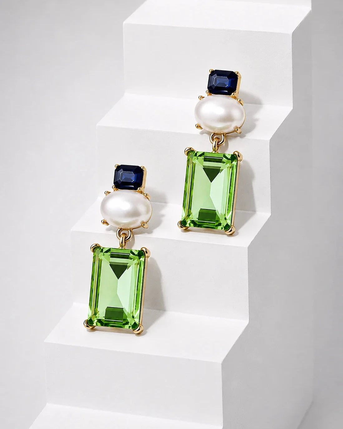 A pair of gold drop earrings featuring rectangular green gemstones, round white pearls, and small dark blue stones, displayed on white geometric steps.