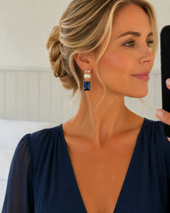 A woman with blonde hair in an updo wearing a dangling earring with a white pearl and a rectangular blue gemstone, holding a phone to take a mirror selfie.