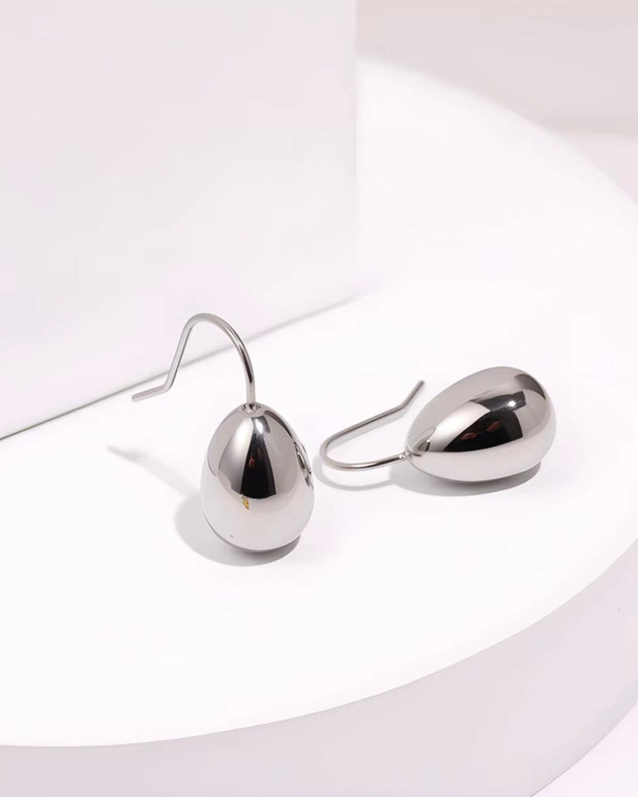 A pair of minimalist silver teardrop-shaped dangle earrings with simple fishhook wires, displayed on a white curved pedestal.