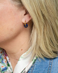 A close-up of a model's ear wearing a small gold hoop earring with a dark blue circular charm, paired with a patterned scarf and denim jacket.